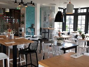 Ein Restaurantinneres mit mehreren Tischen und Stühlen, einige mit Pflanzen. Hängelampen und ein großes Fenster mit Blick.