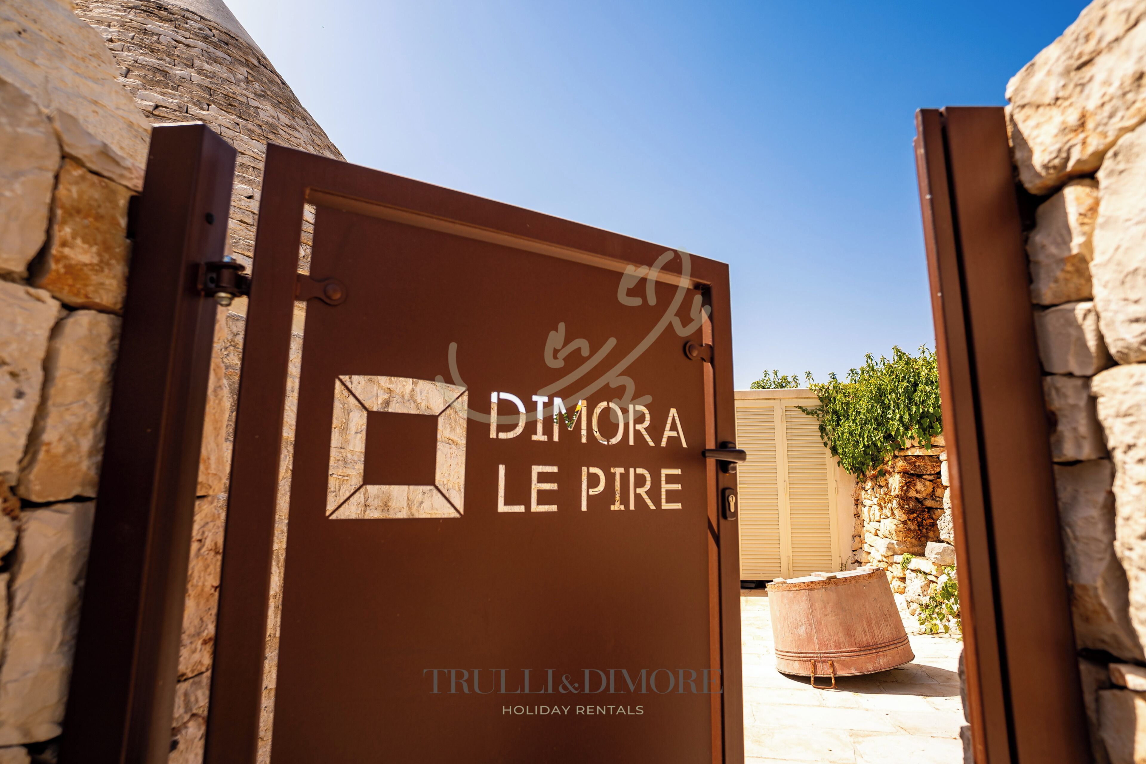 A brown metal gate with the inscription 'DIMORA LE PIRE' and 'TRULLI&DIMORE HOLIDAY RENTALS' indicating a holiday rental property.