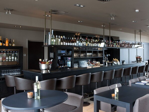 A modern bar area with a long counter, multiple seating options, and a variety of bottles and glasses on display.