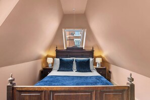 A bedroom with a sloped ceiling, a wooden bed frame, two bedside lamps, and a window with a view of a brick building.
