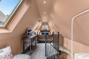 A narrow attic bedroom with a sloped ceiling, a wooden bed, a desk with a chair, a standing fan, and a skylight.
