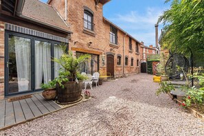 A courtyard with a gravel path, potted plants, outdoor furniture, and a brick building with large windows.