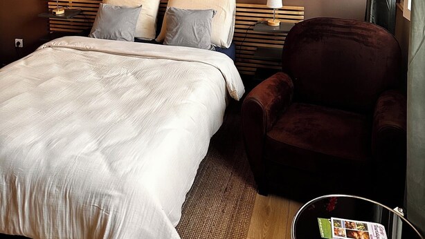 A hotel room with a bed, a chair, a bedside table with a lamp, a vase with flowers, and a painting on the wall.