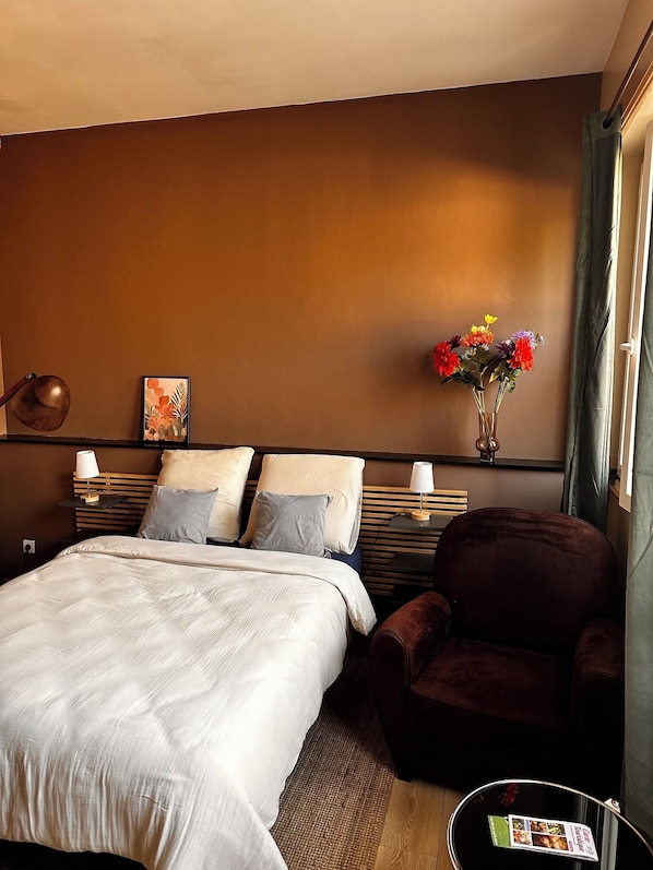 A hotel room with a bed, a chair, a bedside table with a lamp, a vase with flowers, and a painting on the wall.