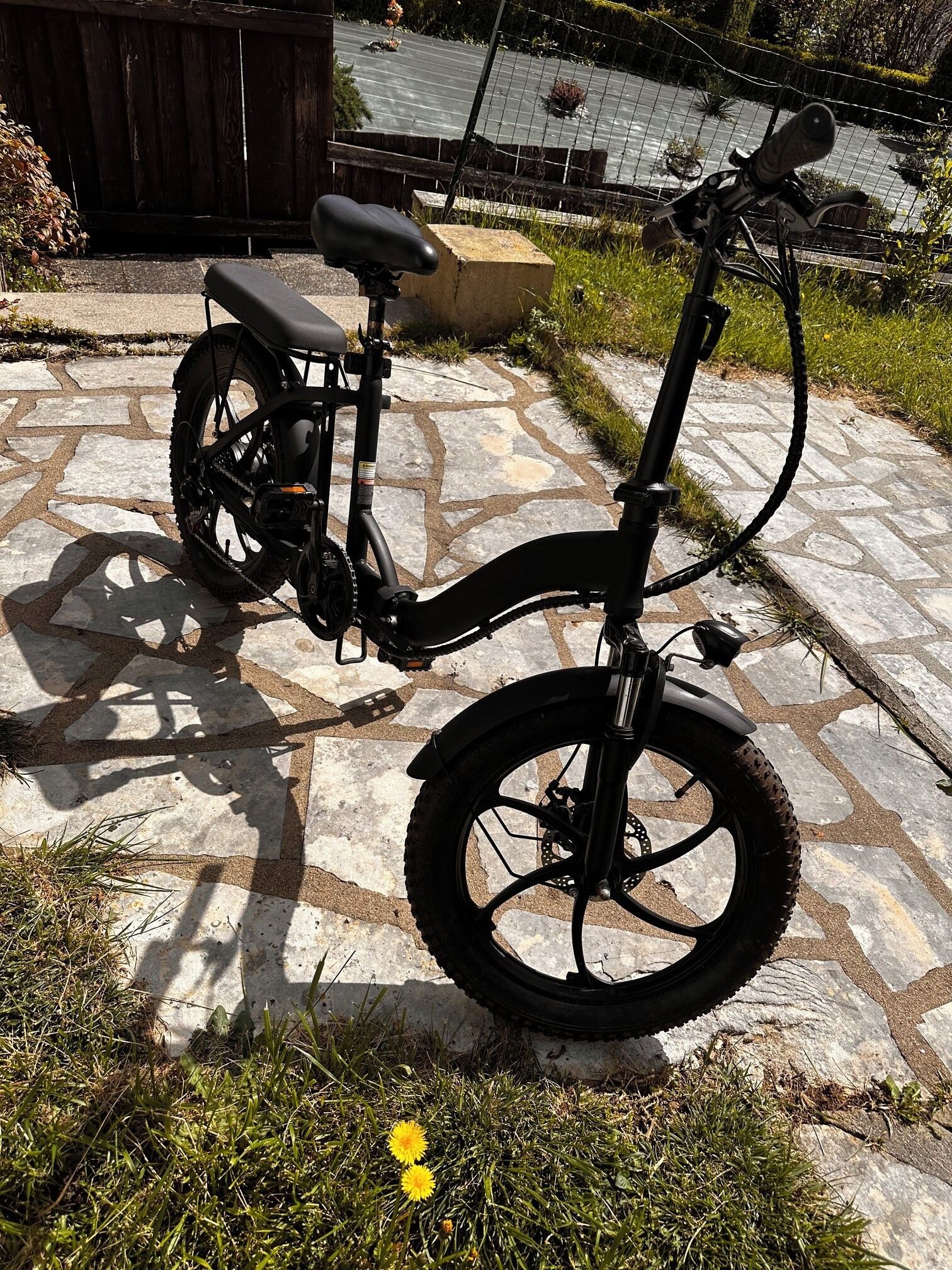 Ein feststehendes E-Bike auf einer Steinterrasse mit einem Holzzaun und einem Wasserspiel im Hintergrund.