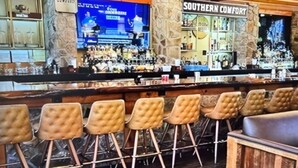 Zona bar con soffitto in legno, bancone e diversi sgabelli imbottiti.