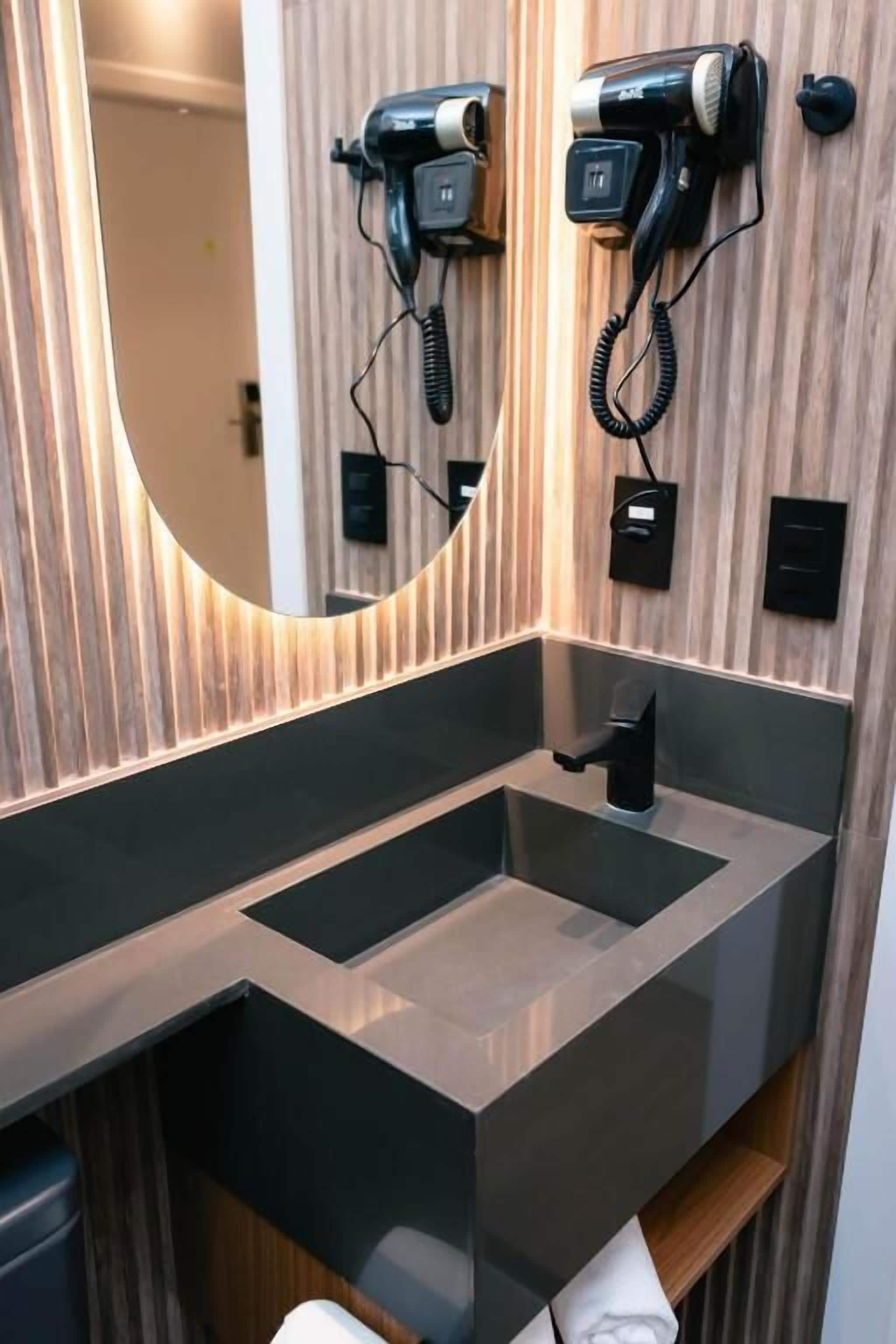 Uma casa de banho moderna com um espelho grande, dois secadores de cabelo fixados na parede e uma lavatório preta elegante.