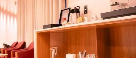 Zona de recepción de hotel con mostrador de madera, pantalla digital y estante rojo con vasos.