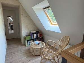 Una habitación con techo inclinado, tragaluz, una silla de mimbre, una estantería y un otoman redondo.