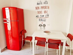 Ein Zimmer mit einem weißen Tisch und roten Stühlen, einem roten Kühlschrank sowie einer Wand mit einem Motivationsspruch und einer Zeichnung von zwei Personen.