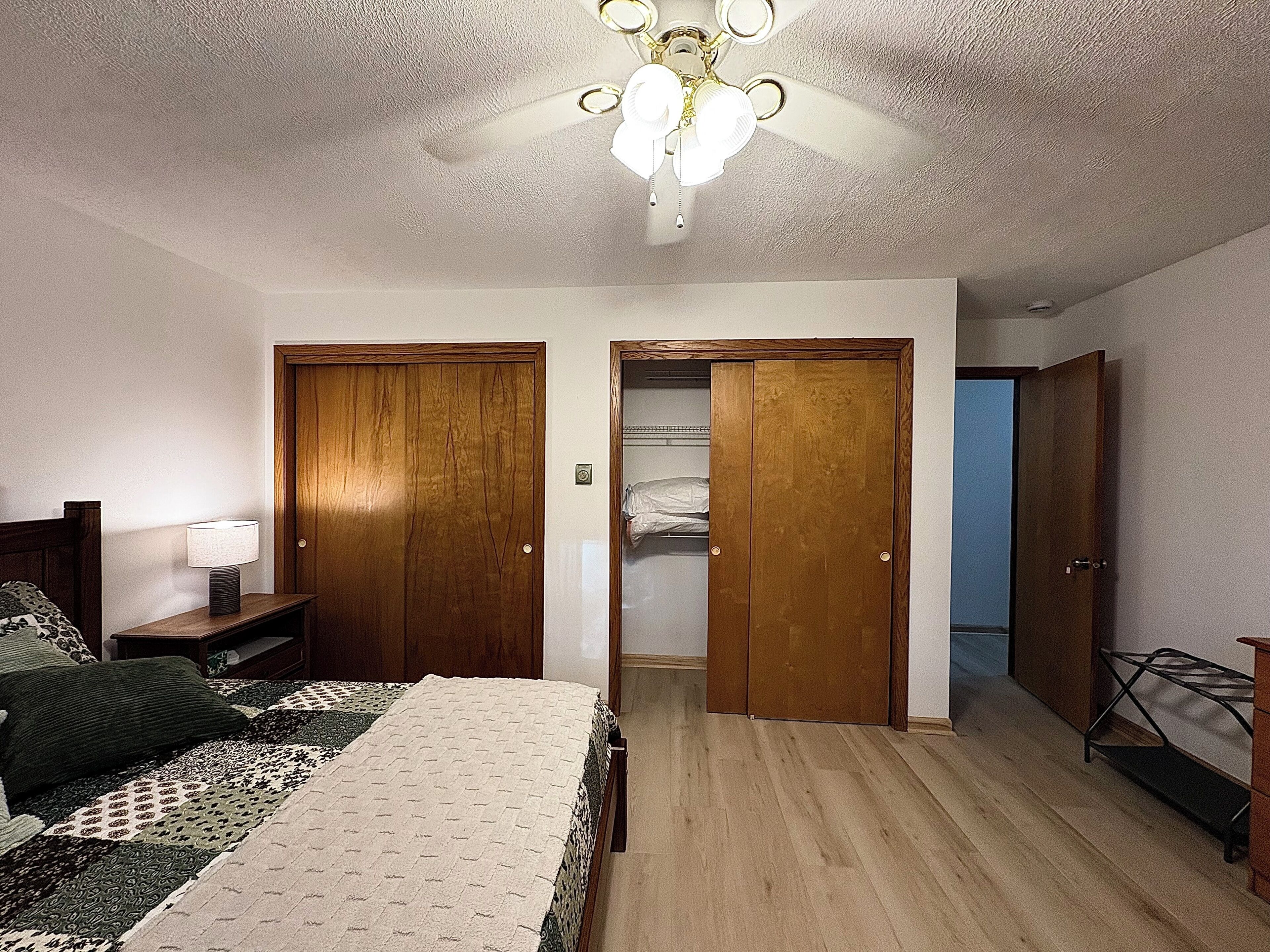 A bedroom with a bed, bedside table, lamp, wooden wardrobe, and a ceiling fan.