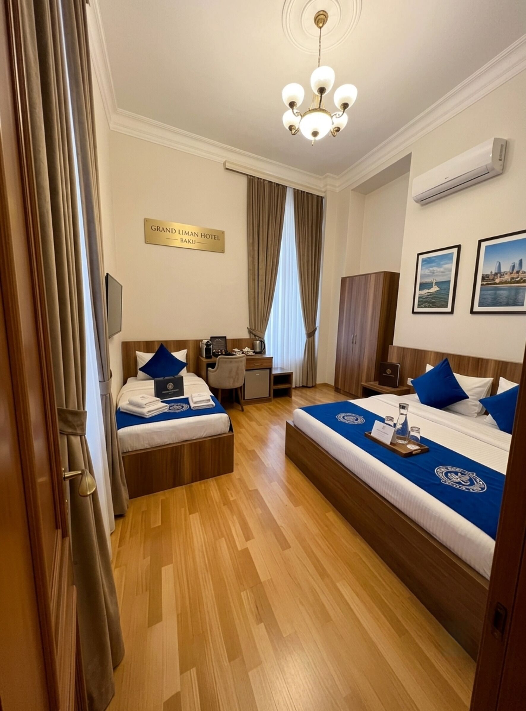 Habitación de hotel con dos camas, un escritorio, un armario, una araña y un letrero con el nombre del hotel.