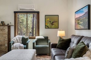 A living room with a fireplace, a large window, a sofa, a chair, a table, and a lamp. - Forest Road Haven: 3 King BR-GameRoom-HotTub-Sauna (Rapid City)