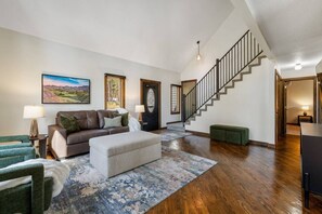 A spacious living room with a brown leather sofa, a matching ottoman, and a large rug. A staircase with a wooden railing leads to an upper level. - Forest Road Haven: 3 King BR-GameRoom-HotTub-Sauna (Rapid City)