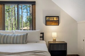 Een slaapkamer met een bed, kussens, een nachtkastje met een lamp, een foto in een lijst aan de muur en een raam met uitzicht op bomen.