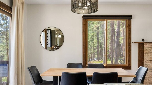 A dining area with a wooden table and black chairs, a round mirror on the wall, and a large window with a forest view. - Forest Road Haven: 3 King BR-GameRoom-HotTub-Sauna (Rapid City)