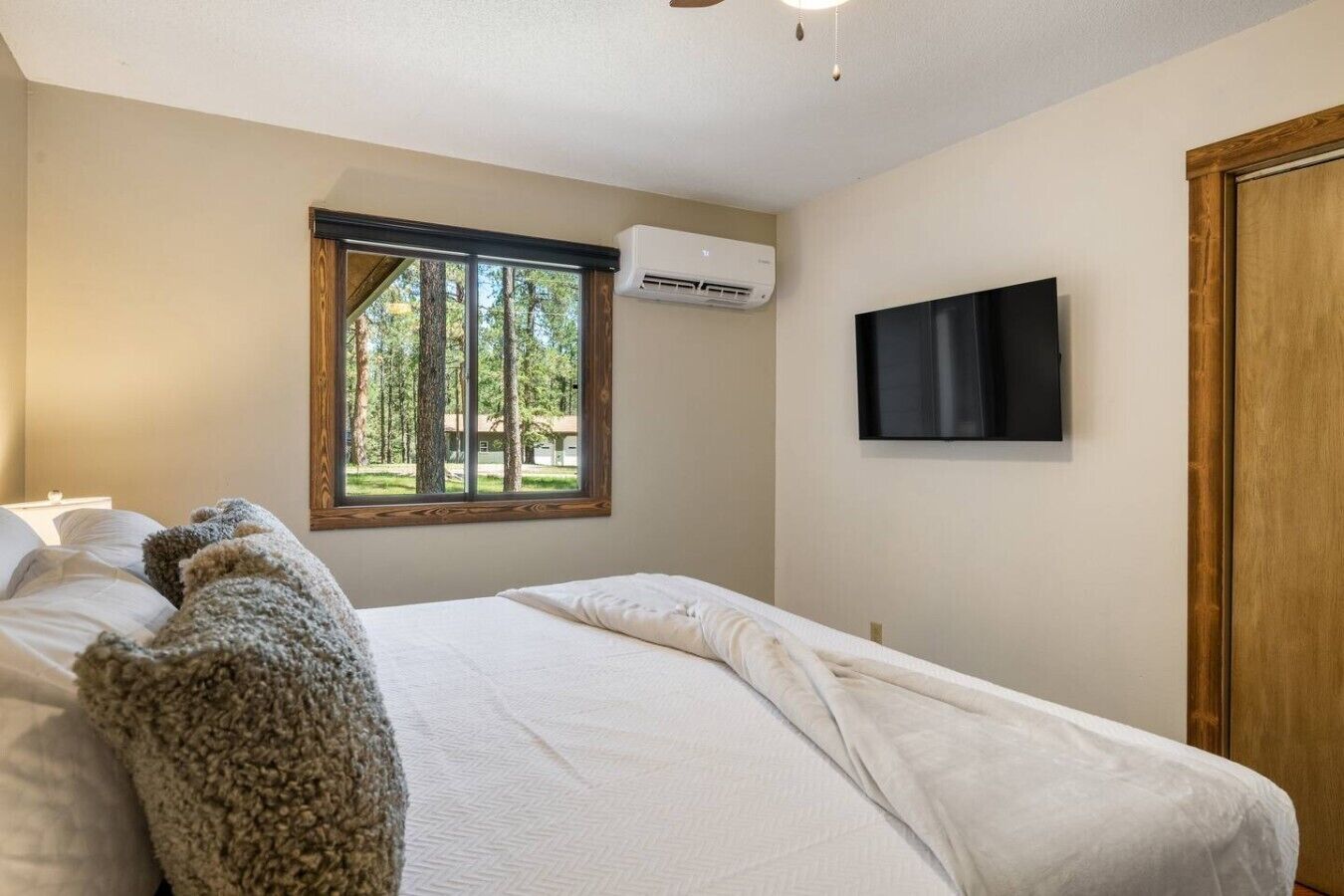 Un dormitorio con cama, ventana con vistas a árboles, unidad de aire acondicionado y televisor montado en la pared.