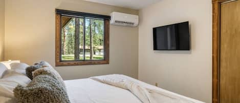 Un dormitorio con cama, ventana con vistas a árboles, unidad de aire acondicionado y televisor montado en la pared.