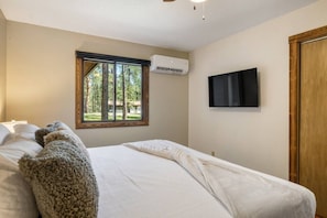 A bedroom with a bed, a window with a view of trees, an air conditioning unit, and a television mounted on the wall. - Forest Road Haven: 3 King BR-GameRoom-HotTub-Sauna (Rapid City)
