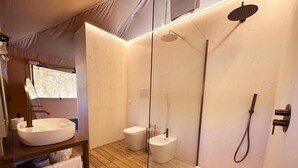 Un baño moderno con piso de madera, un lavamanos blanco grande, un bidé y una zona de ducha con mampara de vidrio.