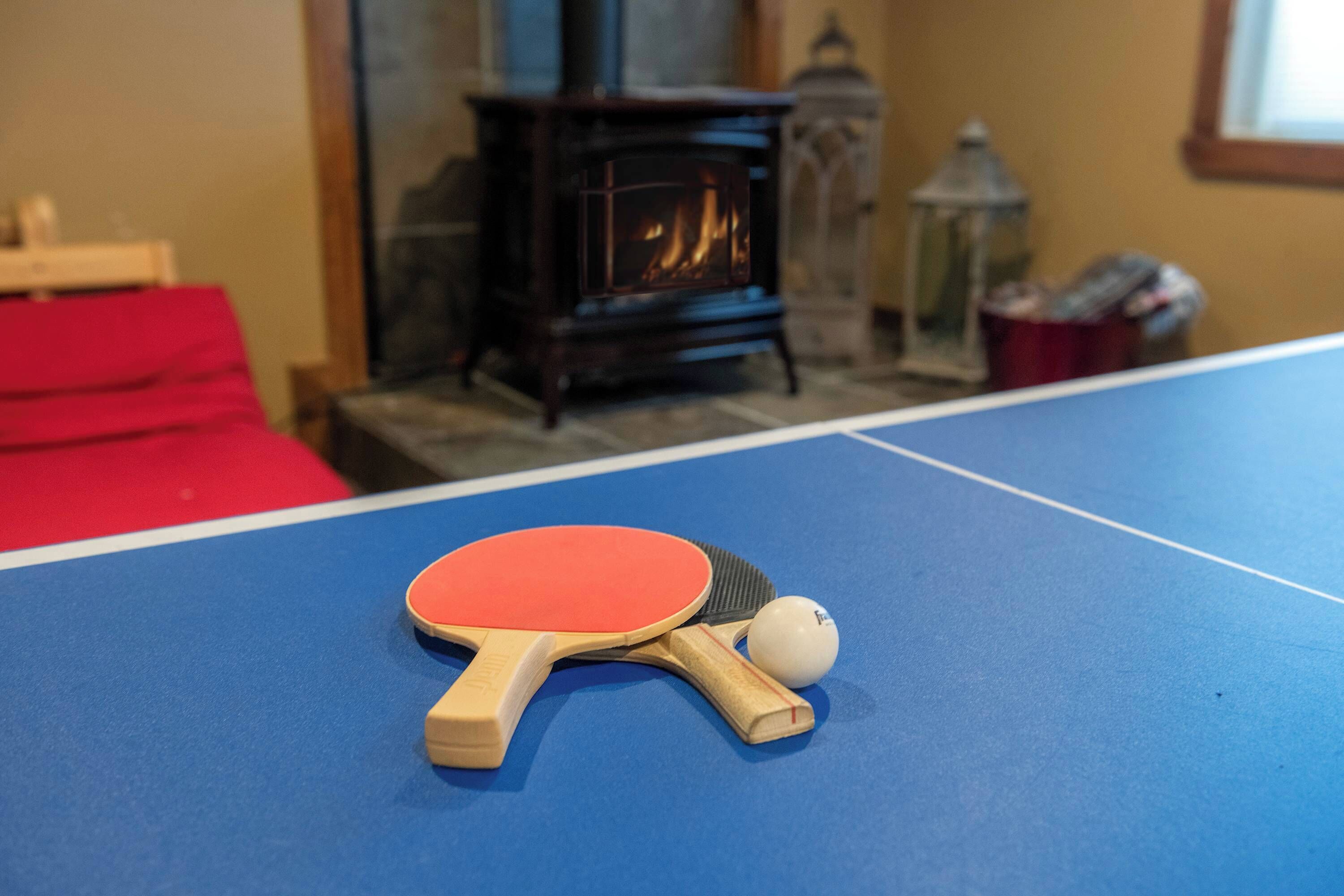 Una mesa de ping pong con dos paletas y una pelota en una habitación con chimenea y un sofá rojo.
