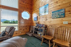 A cozy cabin interior with a leather couch, wooden chairs, a bookshelf, and a large window offering a view of mountains. - Teton Homestead: Bear Creek Cabin Retreat (Driggs)