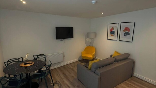 Una sala de estar con un sofá, una mesa de comedor con sillas, un televisor montado en la pared y dos cuadros enmarcados.