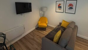 Una sala de estar con un sofá gris, un sillón amarillo y un televisor de pantalla plana montado en la pared.