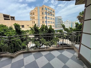 Un balcón con piso a cuadros, baranda de metal y vista de un edificio de varios pisos y ventanas.