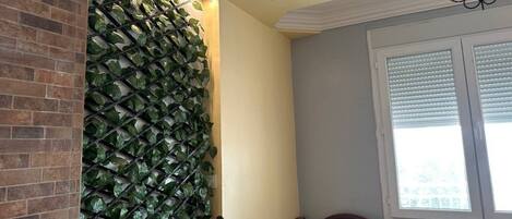 Una habitación con pared de ladrillo, decoración de pared con follaje verde, ventana con persianas y una araña.
