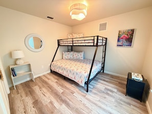A bedroom with a bunk bed, a bedside table with a lamp, a small shelf, a mirror, and a painting on the wall.