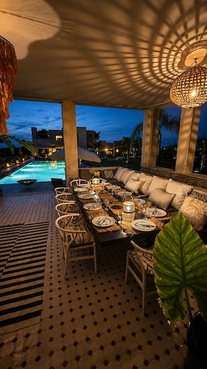 An outdoor dining area with a pool, dining tables set with plates and glasses, and a large cushioned seating area. - "Le Chameau Bleu" Villa: Wellness & Luxury on a 1.5-hectare private estate (Marrakech)