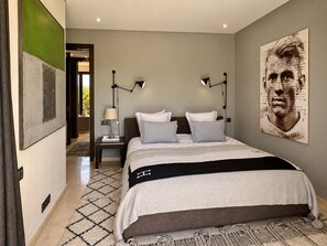 A bedroom with a bed, two bedside tables, a wall-mounted lamp, a large framed picture, and a patterned rug. - "Le Chameau Bleu" Villa: Wellness & Luxury on a 1.5-hectare private estate (Marrakech)