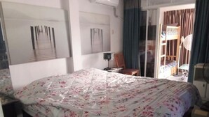 A hotel room with a bed, a desk, a chair, a window with curtains, and a bunk bed in the background. - Room-big Charming Suite With Balcony in (Pattaya)