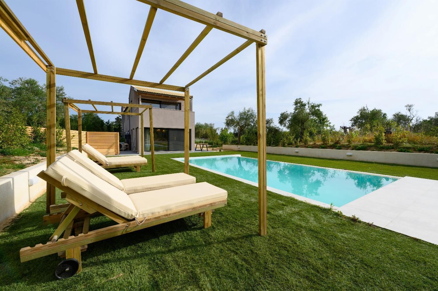 Een zwembadgedeelte met een houten schommelstoel en ligstoelen, een modern huis op de achtergrond, en een verzorgd gazon.