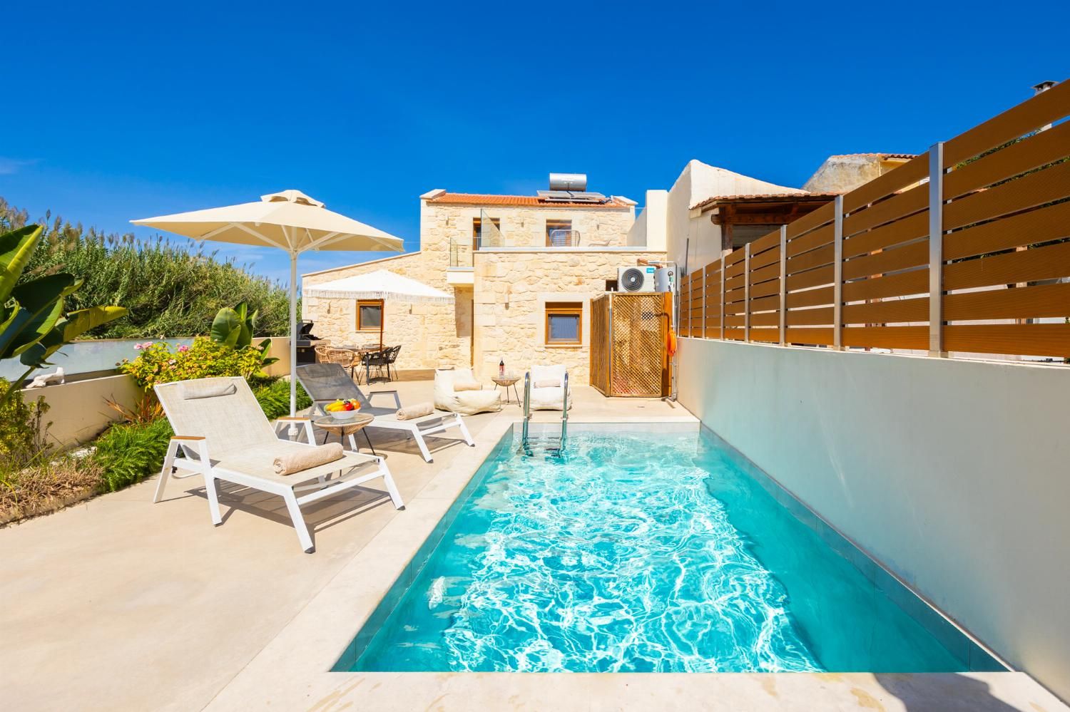 A swimming pool with lounge chairs and umbrellas, a stone building, and a wooden fence.