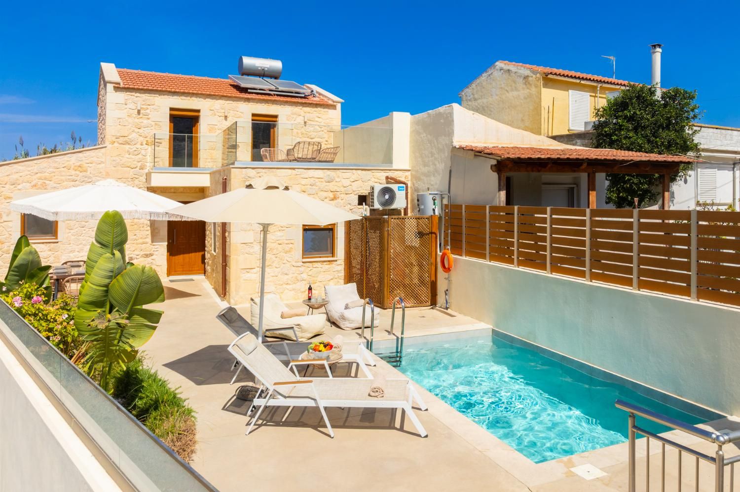 Un espace aménagé au bord de la piscine, avec des chaises longues, un coin salon abrité et un bâtiment en pierre surmonté d’un toit en tuiles.
