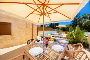 Área de refeições ao ar livre com mesa posta para dois, um guarda-sol grande e piscina ao fundo.