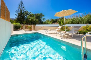 Ein Schwimmbecken mit Liegestühlen und einem Sonnenschirm, umgeben von Grünpflanzen und blauem Himmel.