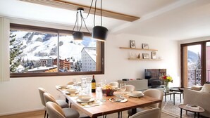 Ein moderner Essbereich mit großem Fenster mit Blick auf eine verschneite Berglandschaft, ein Holztisch gedeckt für eine Mahlzeit und eine gemütliche Sitzecke mit Flachbildfernseher.