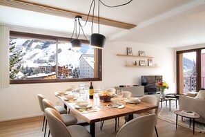 A modern dining area with a large window overlooking a snowy mountain landscape, a wooden dining table set for a meal, and a cozy seating area with a flat-screen TV. - Super Venosc | T3 à 100m des pistes, 50m télésiège (MONT-DE-LANS)