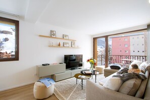 A modern living room with a television, sofa, and a balcony with a view of a building. - Super Venosc | T3 à 100m des pistes, 50m télésiège (MONT-DE-LANS)