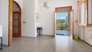 A spacious room with a tiled floor, a wooden door, a small table with a lamp, and a view through an open doorway. - Apartment 'Alle Colonne Bianche Rooms' with Balcony, Wi-Fi and Air Conditioning (Conversano)