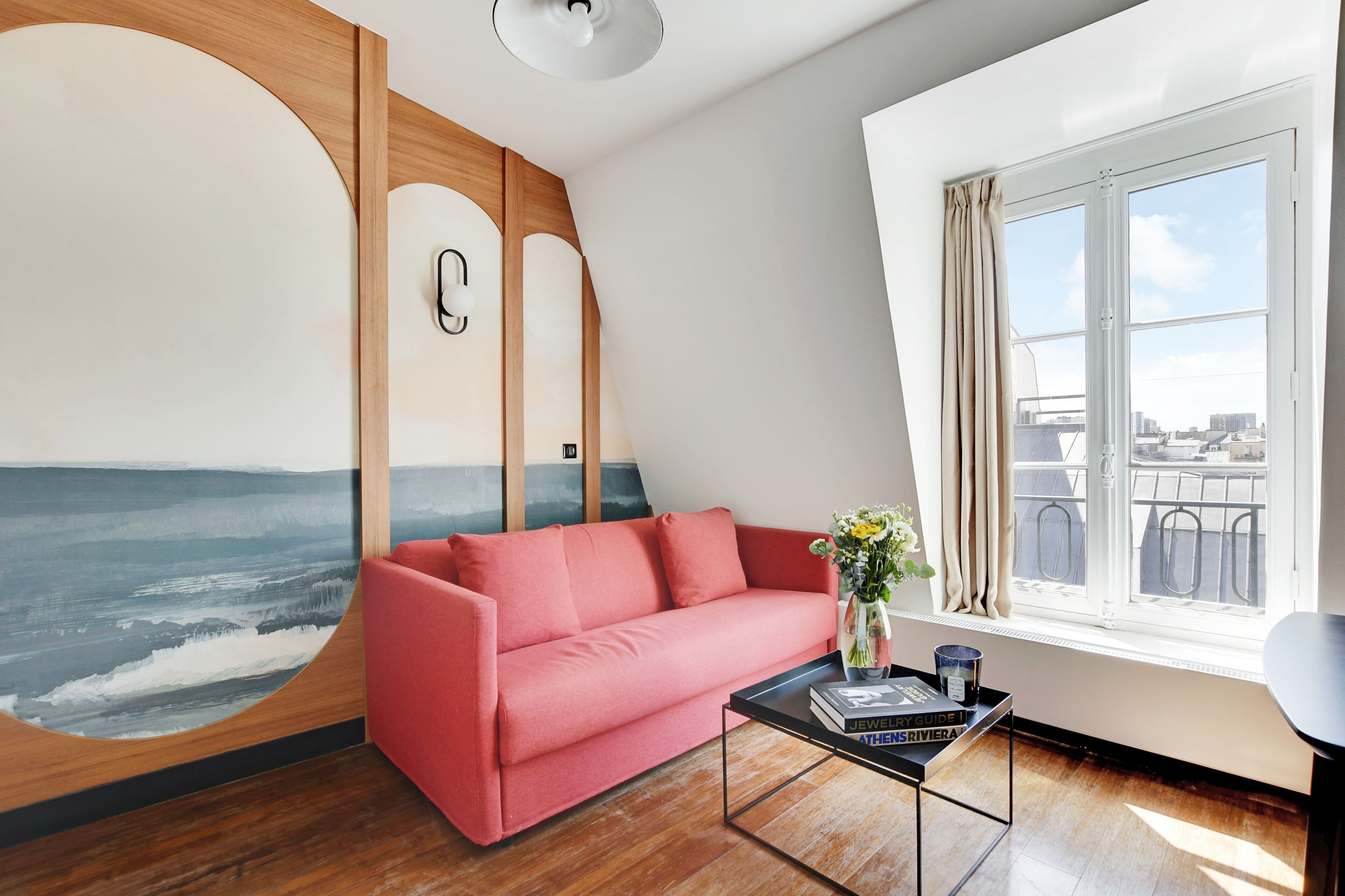 Uma sala de estar moderna com um sofá vermelho, uma mesa de centro com livros, um mural na parede e uma janela com vista para a cidade.
