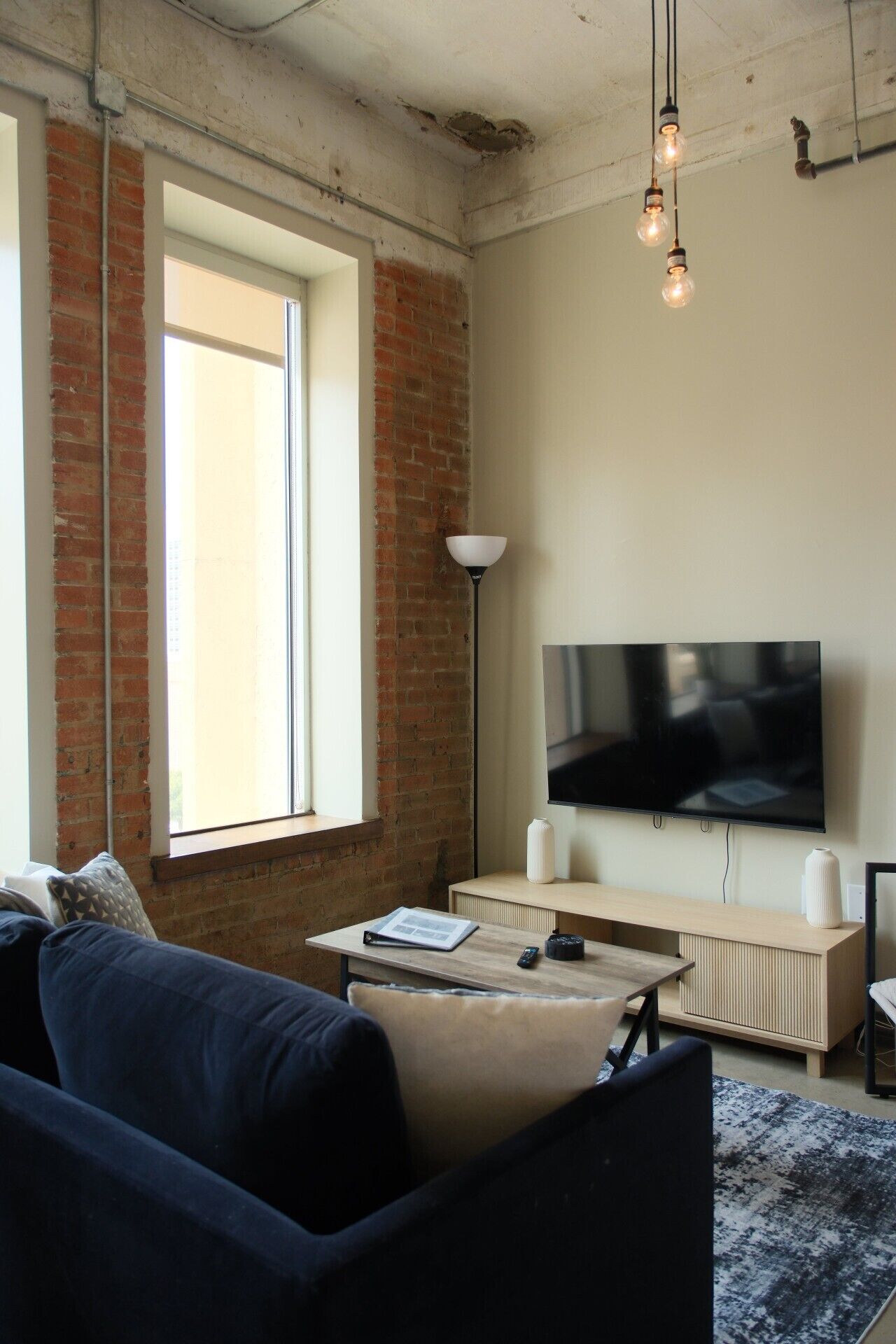 A modern living room with a brick wall, a large window, a flat-screen TV, a sofa, a coffee table, and a floor lamp.