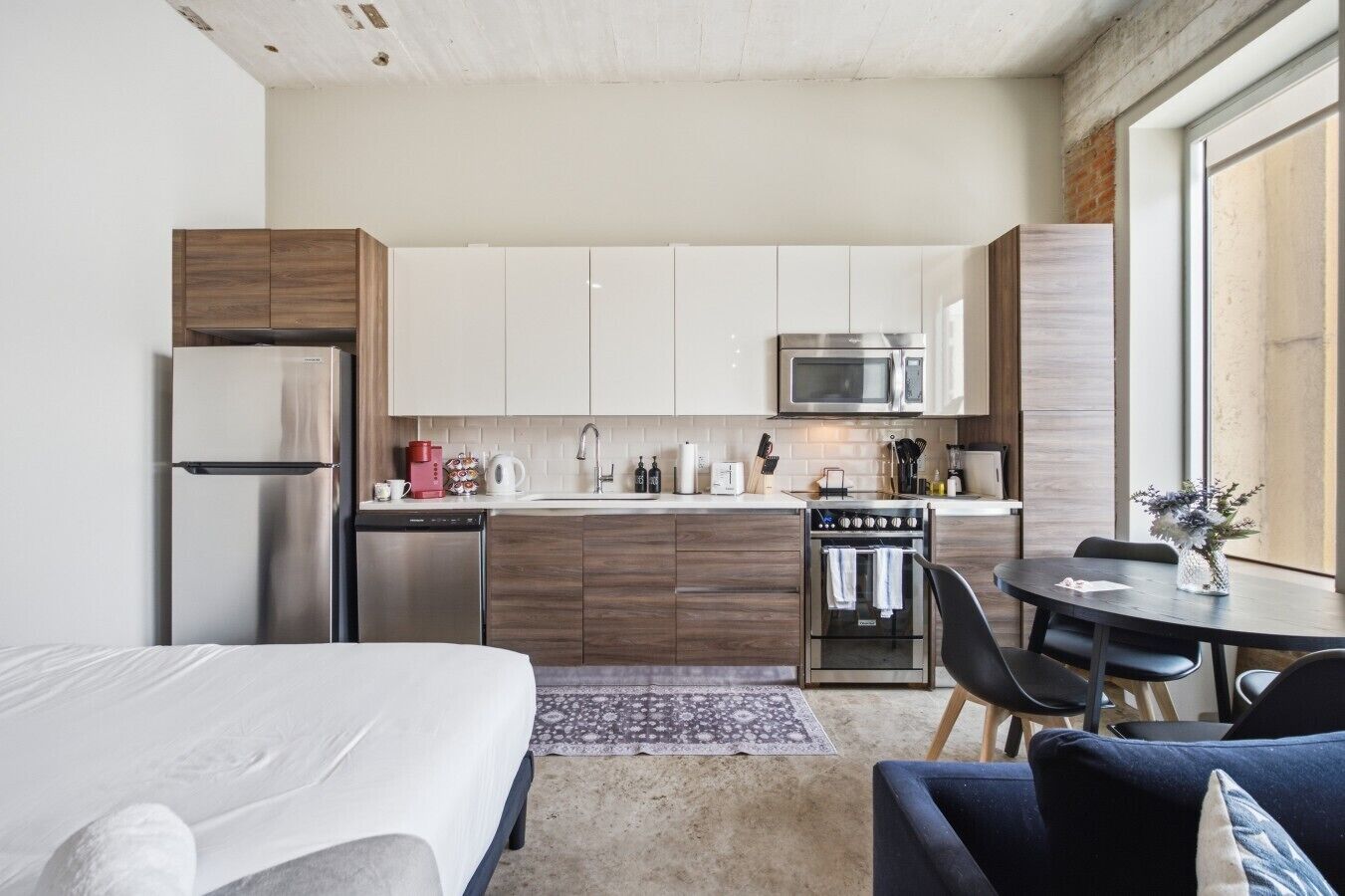 A compact kitchen with a refrigerator, microwave, and oven, a dining table with chairs, and a bed with a patterned rug.