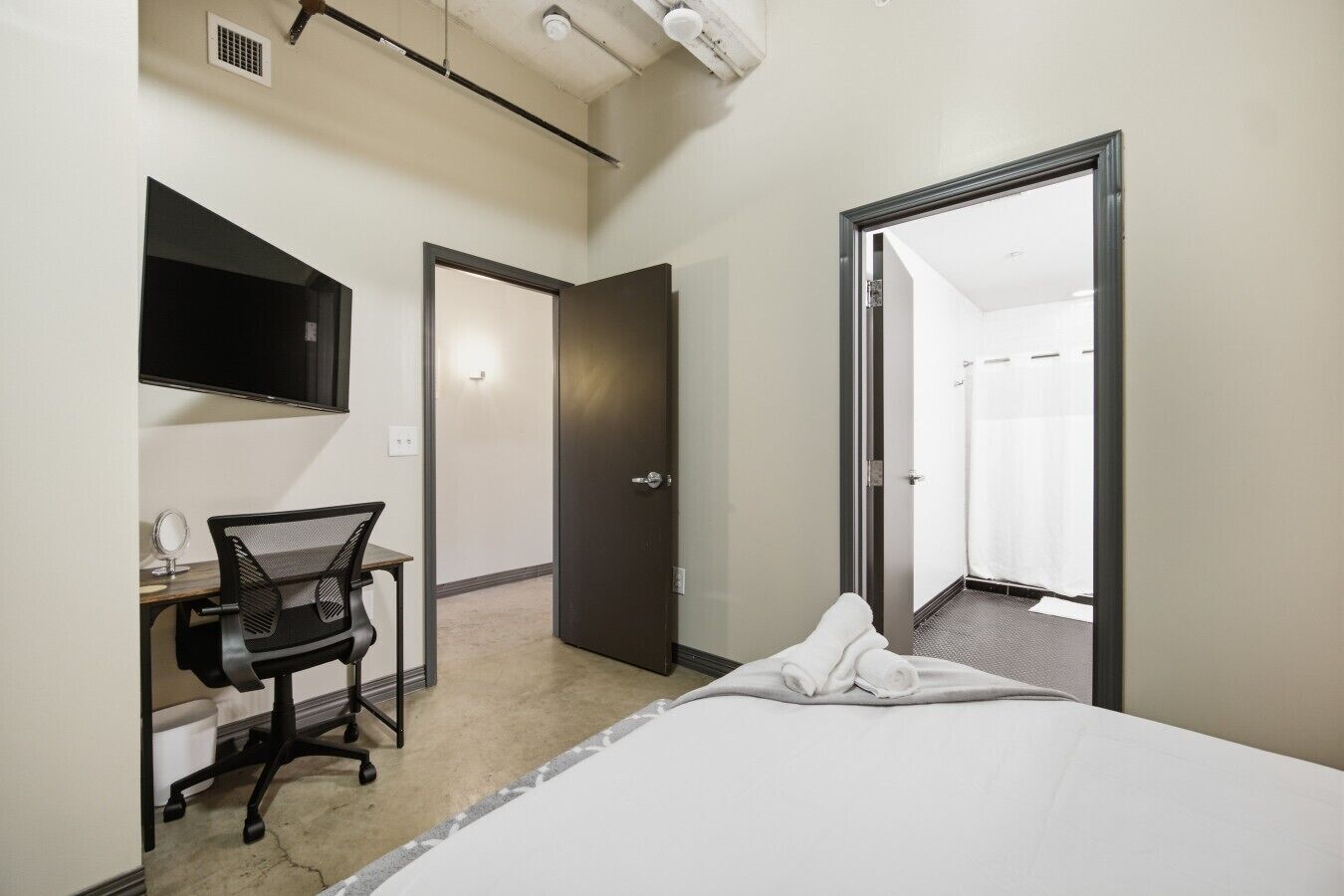 A hotel room with a bed, a desk with a chair, a mirror, a television, and a door leading to a bathroom.