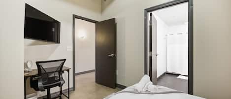 Habitación de hotel con cama, escritorio con silla, espejo, televisión y puerta que da a un baño.