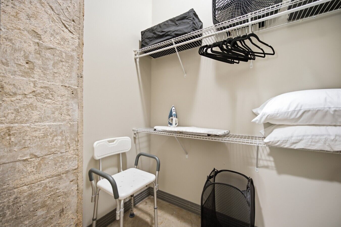 Habitación pequeña y funcional con estructura metálica para la cama, una silla, un estante con una botella de agua y una bolsa de malla negra.