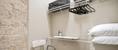 Habitación pequeña y funcional con estructura metálica para la cama, una silla, un estante con una botella de agua y una bolsa de malla negra.